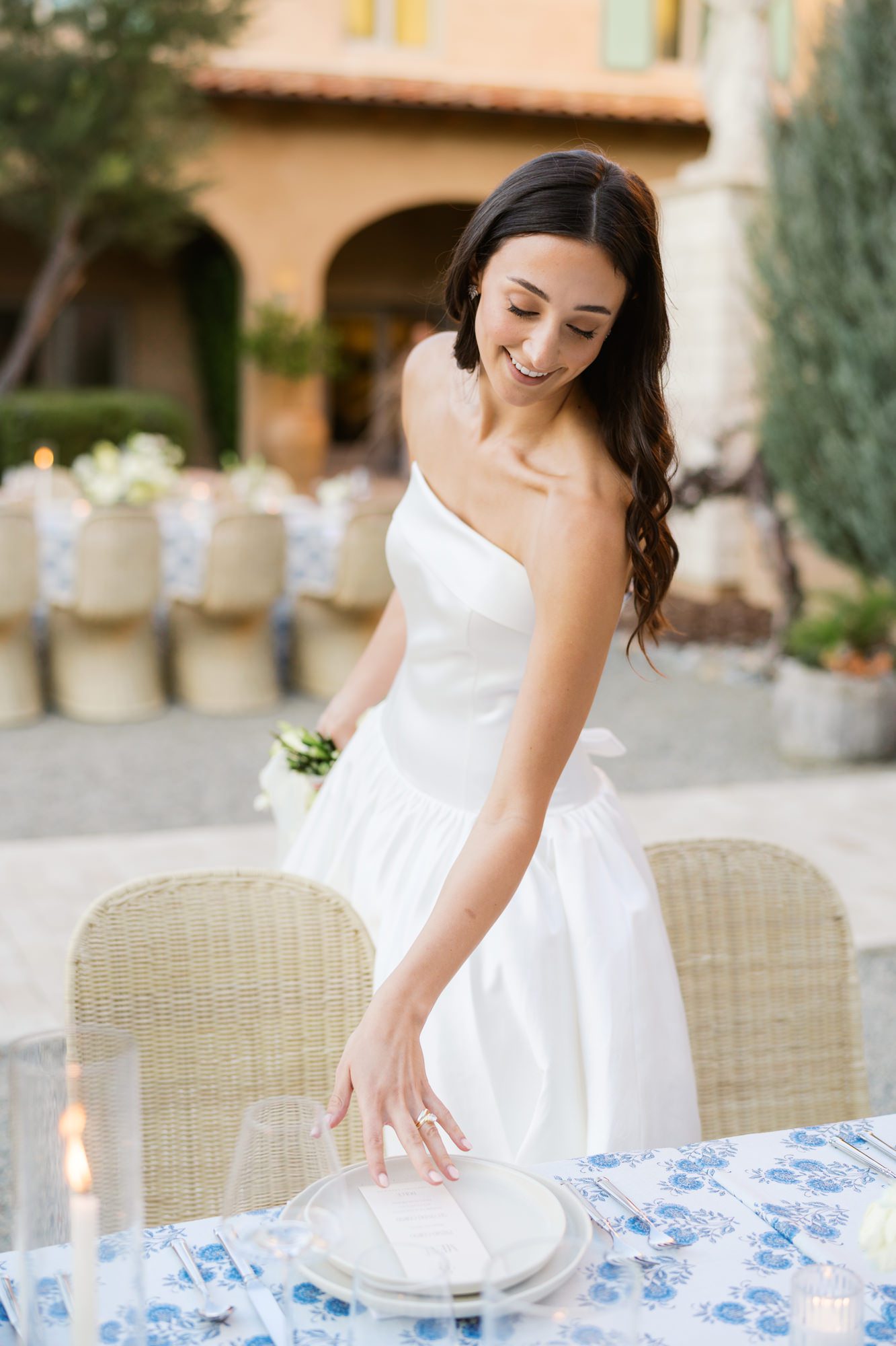Bride setting a reception table with woven chairs and blue floral table linen in a sun-washed courtyard setting.
