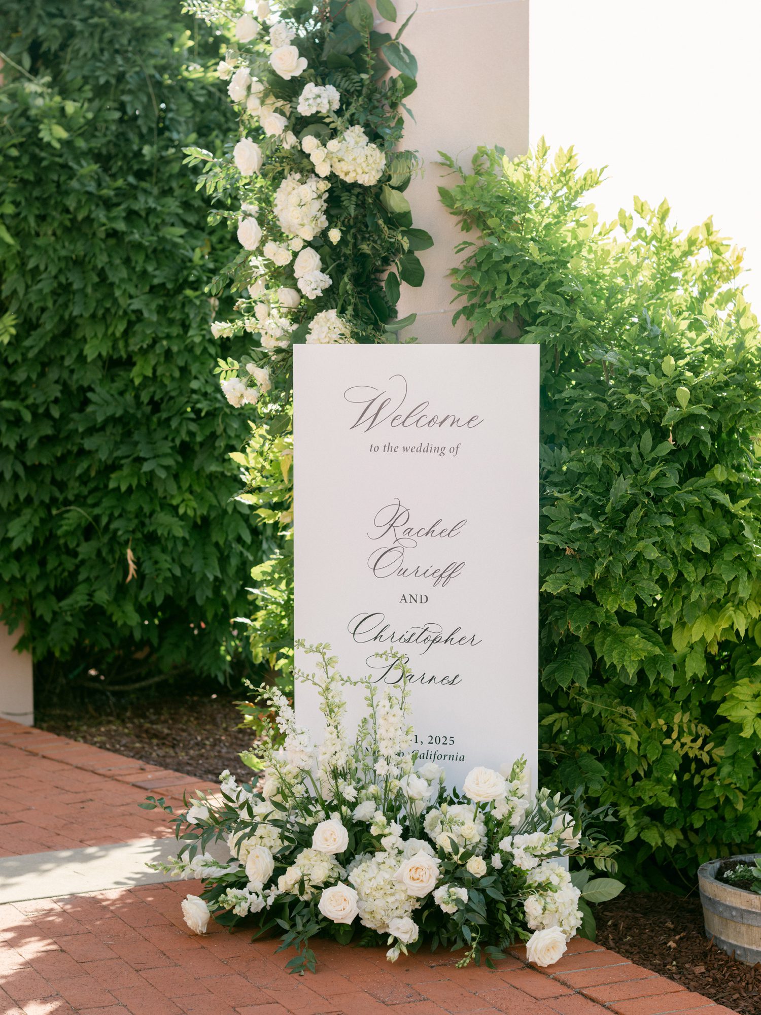 Elegant white wedding welcome sign with script typography and white floral arrangement in garden setting