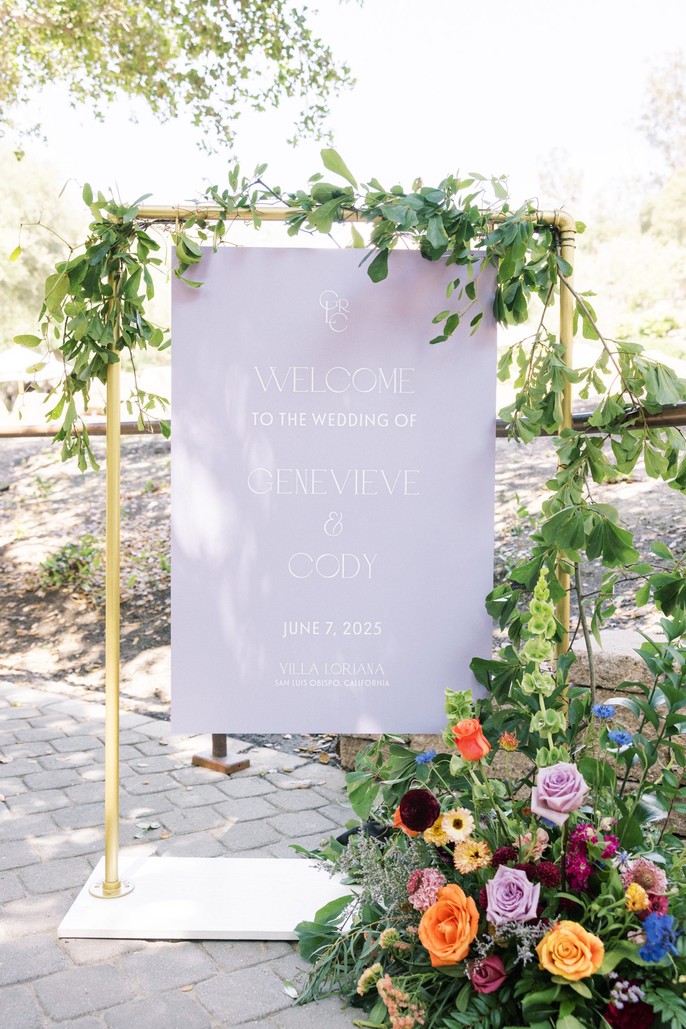Lavender wedding welcome sign with white text framed in brass with garden florals
