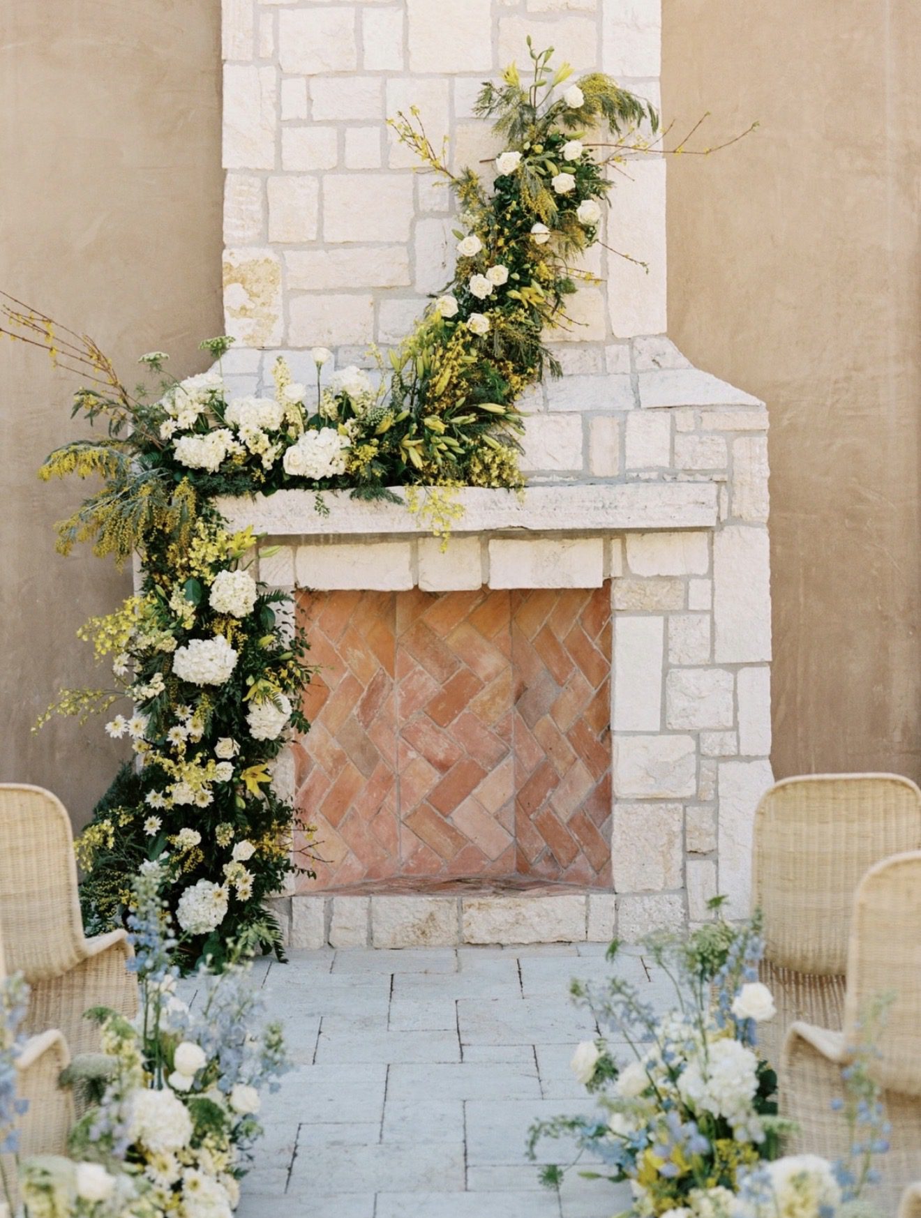 Stone fireplace adorned with soft white and yellow florals in an Italian-inspired courtyard wedding setting.