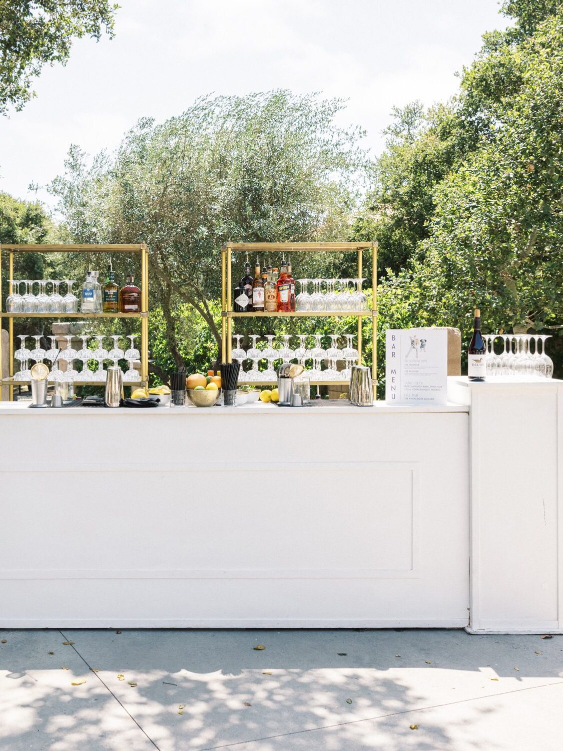 Outdoor wedding bar setup with glassware, bottles, and gold shelving